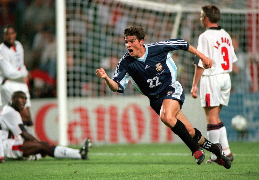 Futbolista de la Selección argentina celebra un gol contra Inglaterra en un partido clave, con jugadores rivales en el suelo.