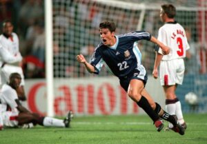 Futbolista de la Selección argentina celebra un gol contra Inglaterra en un partido clave, con jugadores rivales en el suelo.